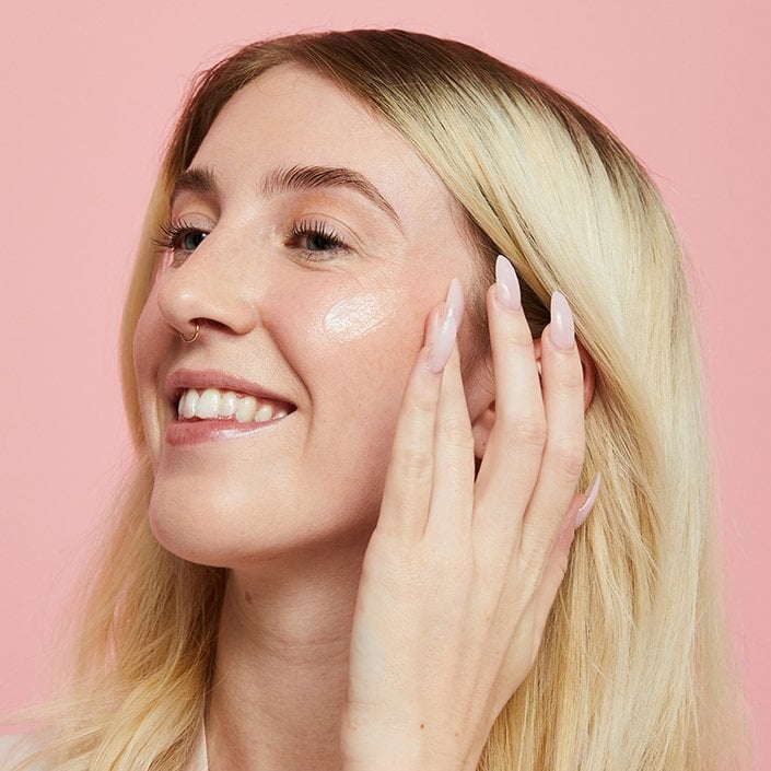 Picture of a blonde person smiling in profile and applying moisturizer to their cheek
